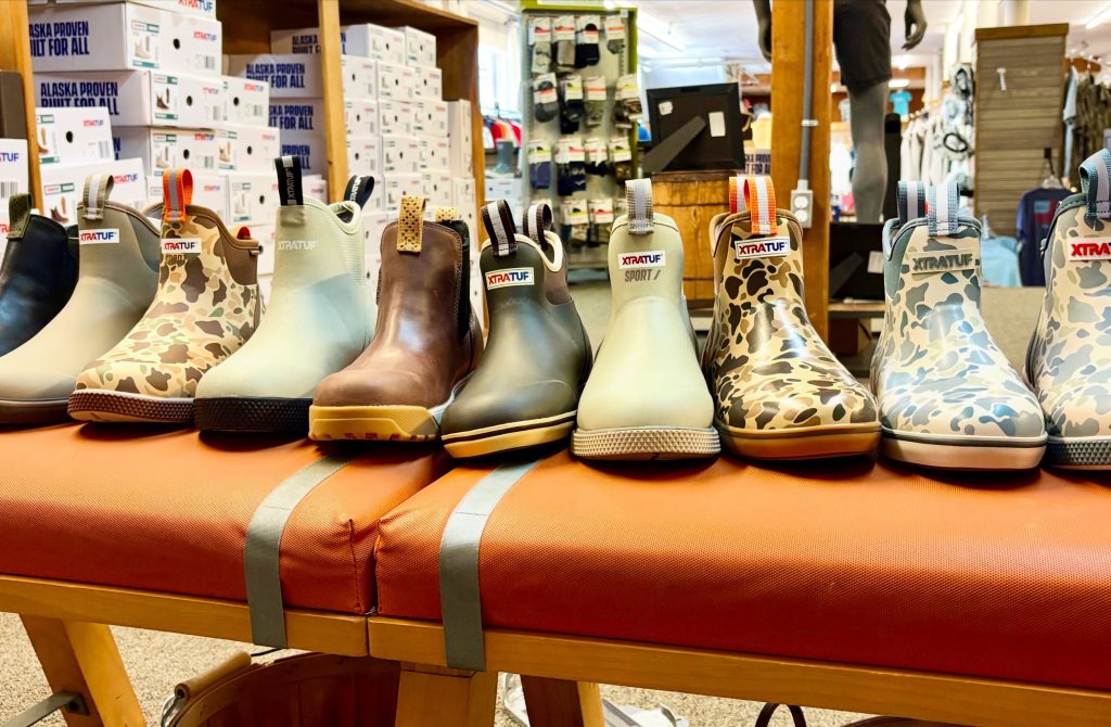 Row of camouflage-patterned XTRATUF rain boots on a wooden display in a store window.