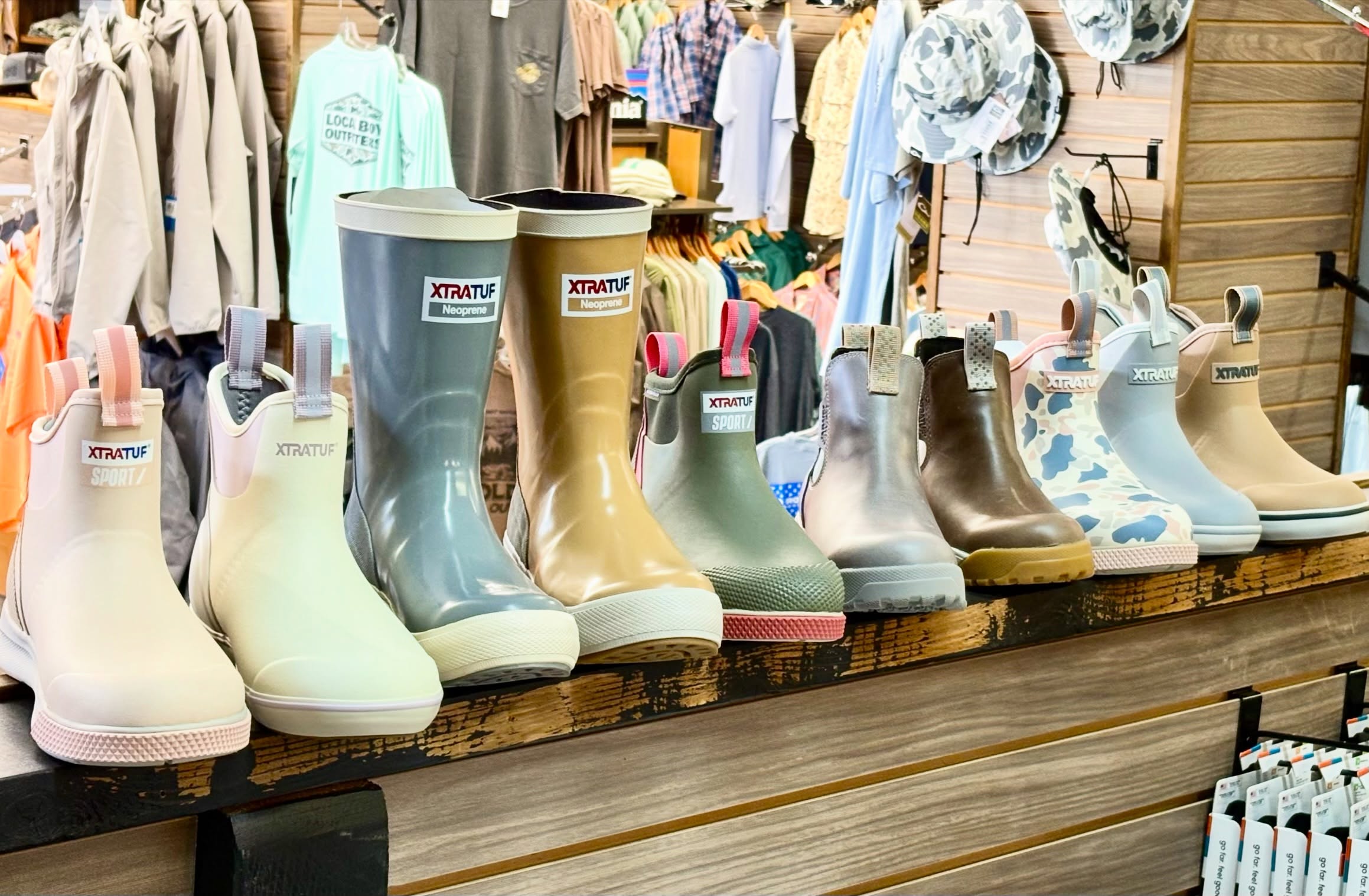 Row of XTRATUF rain boots in green, gray, and camo on a rustic wooden display shelf in a clothing store, background with jackets.
