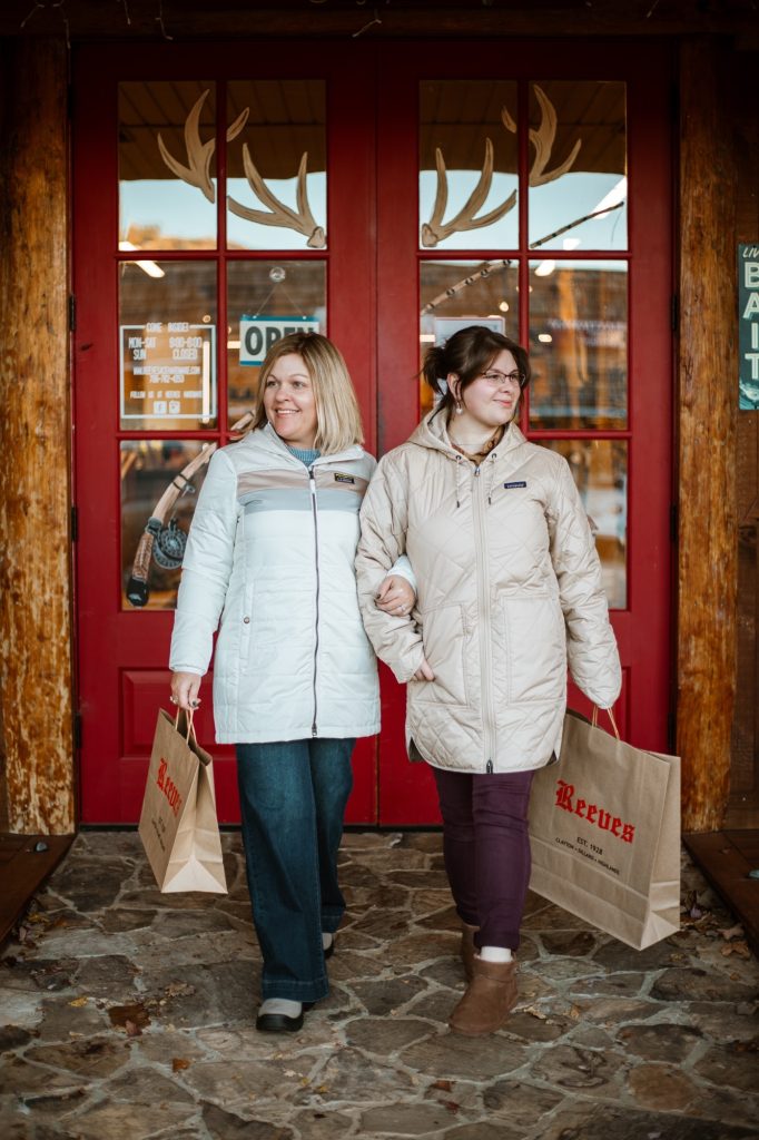 5Y8A0833-2 - Reeves 1928 Two women in light jackets walk out of a shop, carrying Reeves paper bags, in front of a red door with antler decor.