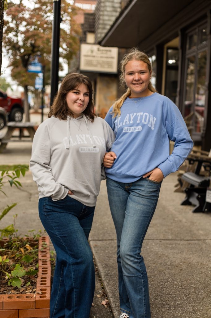 5Y8A3655 - Reeves 1928 Two young women stand arm-in-arm on a city sidewalk, wearing Clayton Georgia sweatshirts and smiling at the camera.