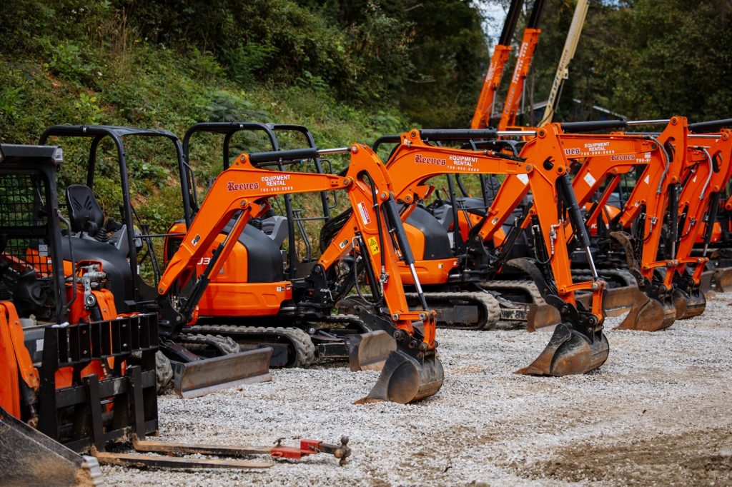Row of bright orange construction excavators parked on a gravel lot with green bushes in the background for scale and context.