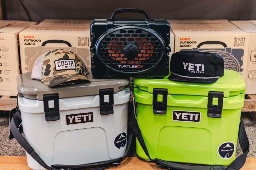 5Y8A6674 - Reeves 1928 Two YETI coolers (gray and lime green) sit side by side with a portable speaker on top; camouflage cap and boxed outdoor gear behind them.