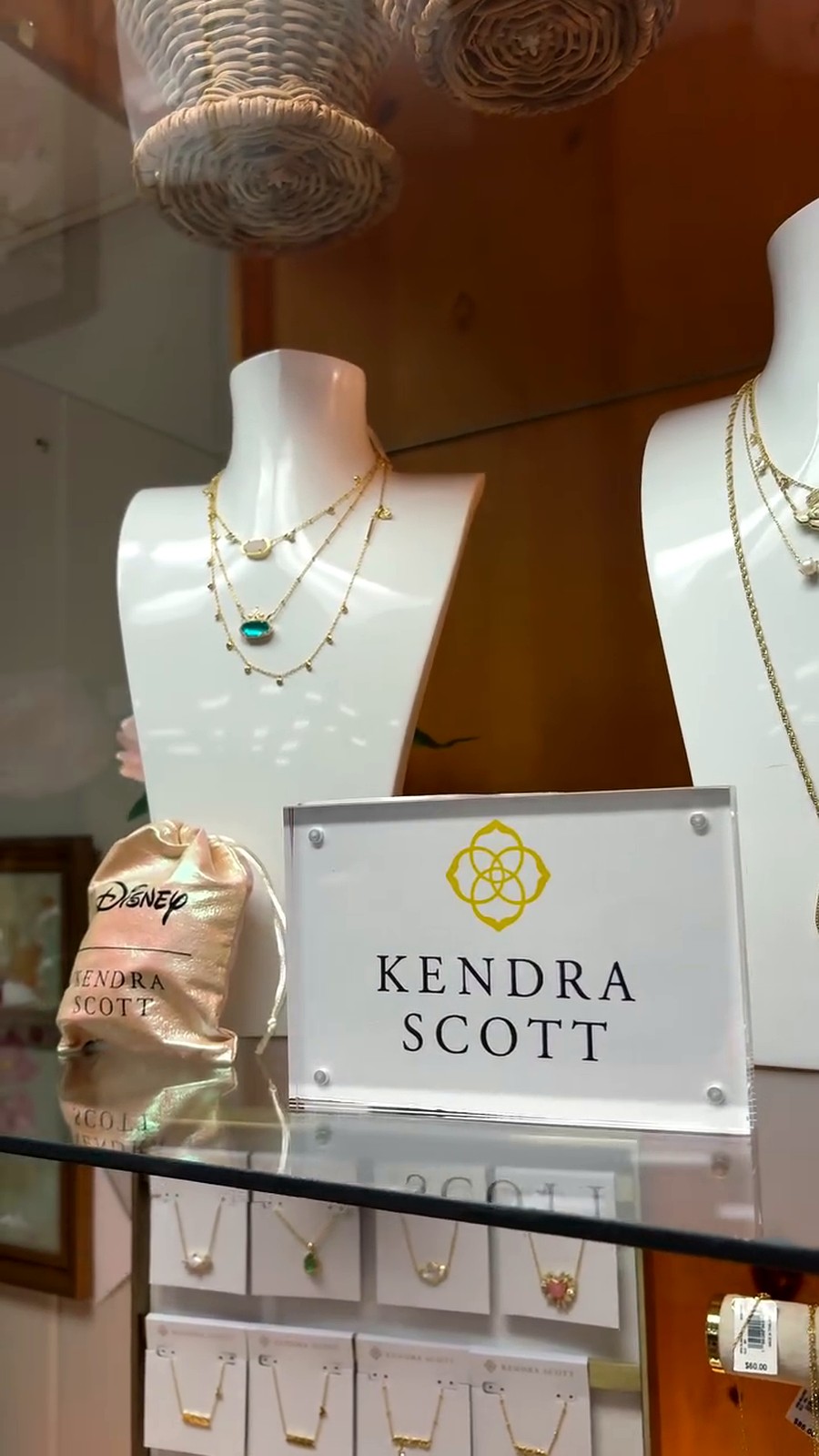 Two white display busts showcase layered gold necklaces, one with turquoise stone, on a jewelry shelf with a Kendra Scott sign nearby.