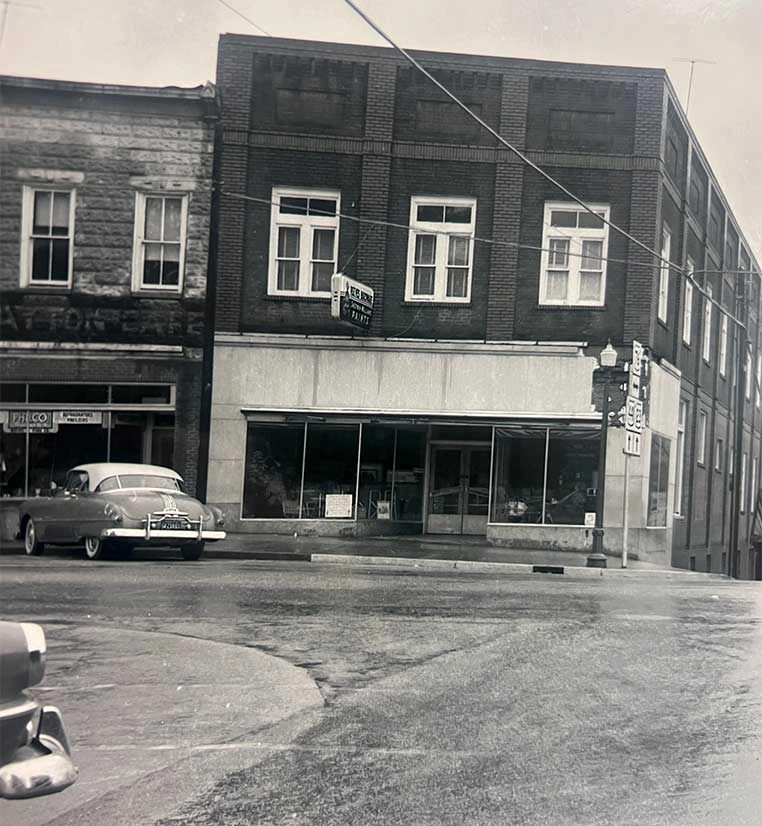 Black-and-white street scene with a two-story brick storefront and large display windows; a vintage car is parked along the curb in front.