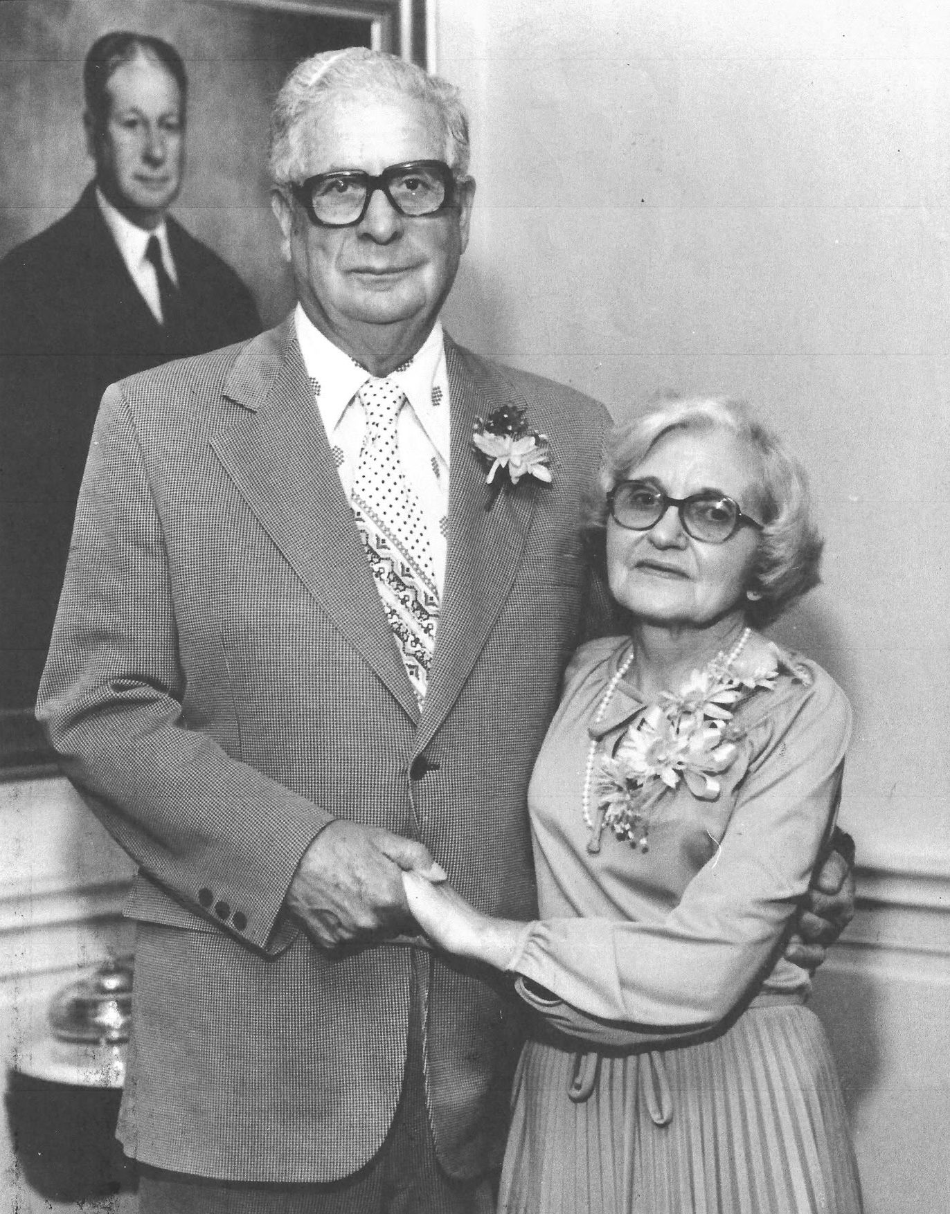 Older couple at a formal event; man in a plaid suit with boutonniere and patterned tie, woman in a striped dress with a corsage and pearls, both wearing glasses, holding hands.