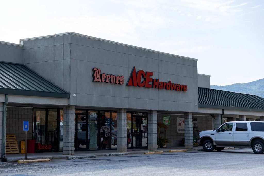 5Y8A8441 - Reeves 1928 Front of an ACE Hardware store with a red sign on a gray building, glass doors, and a white pickup parked out front.