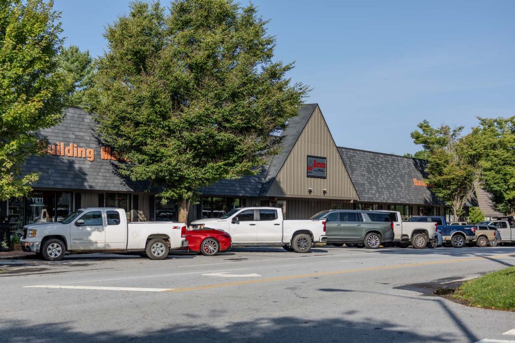 Strip mall with a tall tree in front and several pickup trucks parked along the curb on a sunny day in front of the shops.
