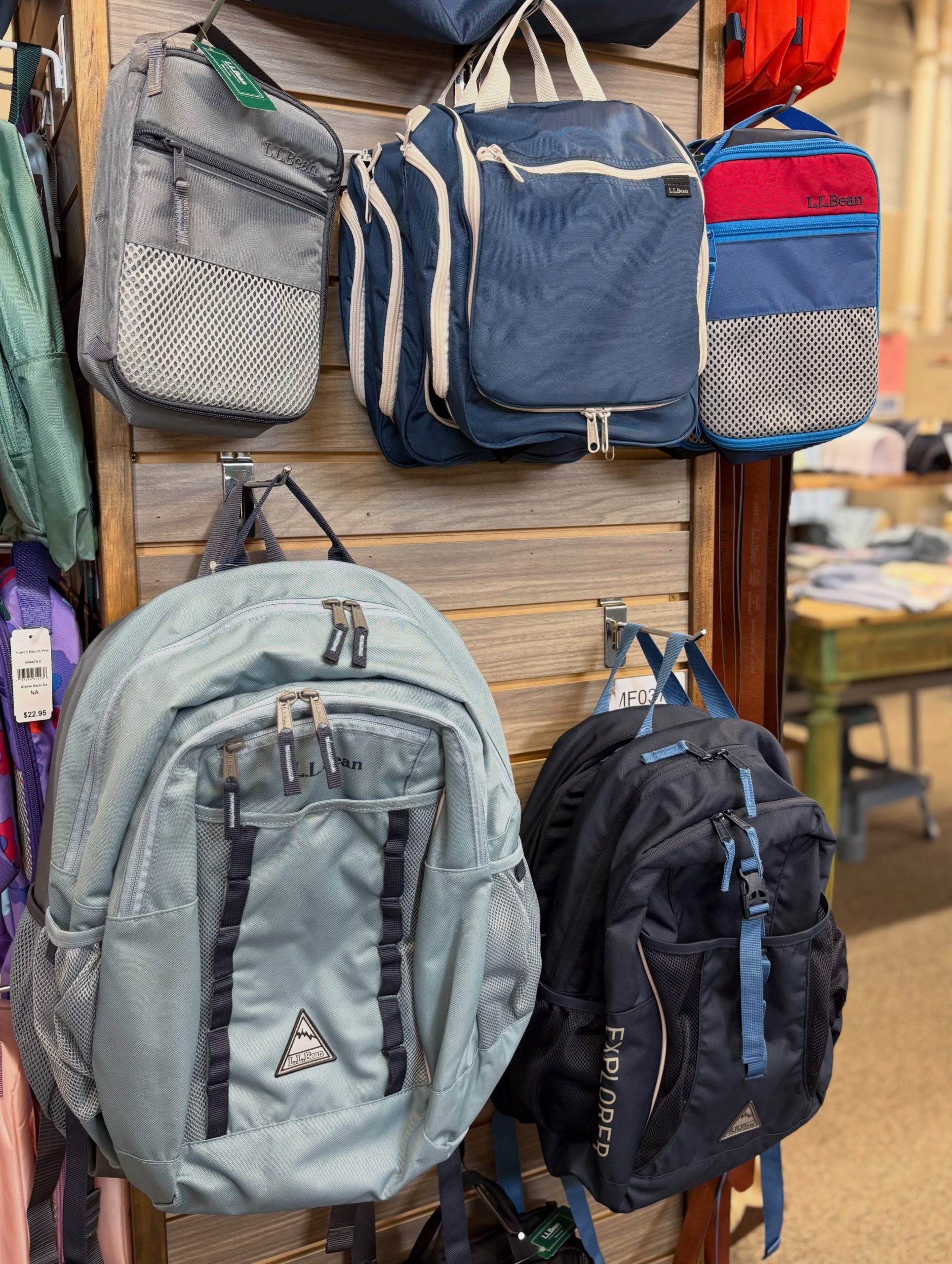 Backpacks hanging on a wooden rack in a store; gray, blue, and red bags with mesh pockets and LL Bean branding.