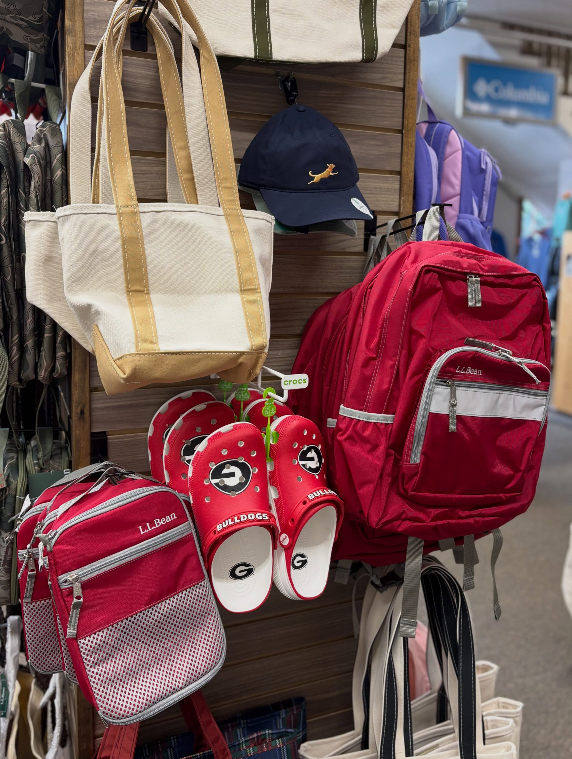Retail display of bags and accessories: beige tote with tan straps, navy cap with embroidered dog, and red LL Bean backpacks with a pair of red Bulldog slippers nearby.