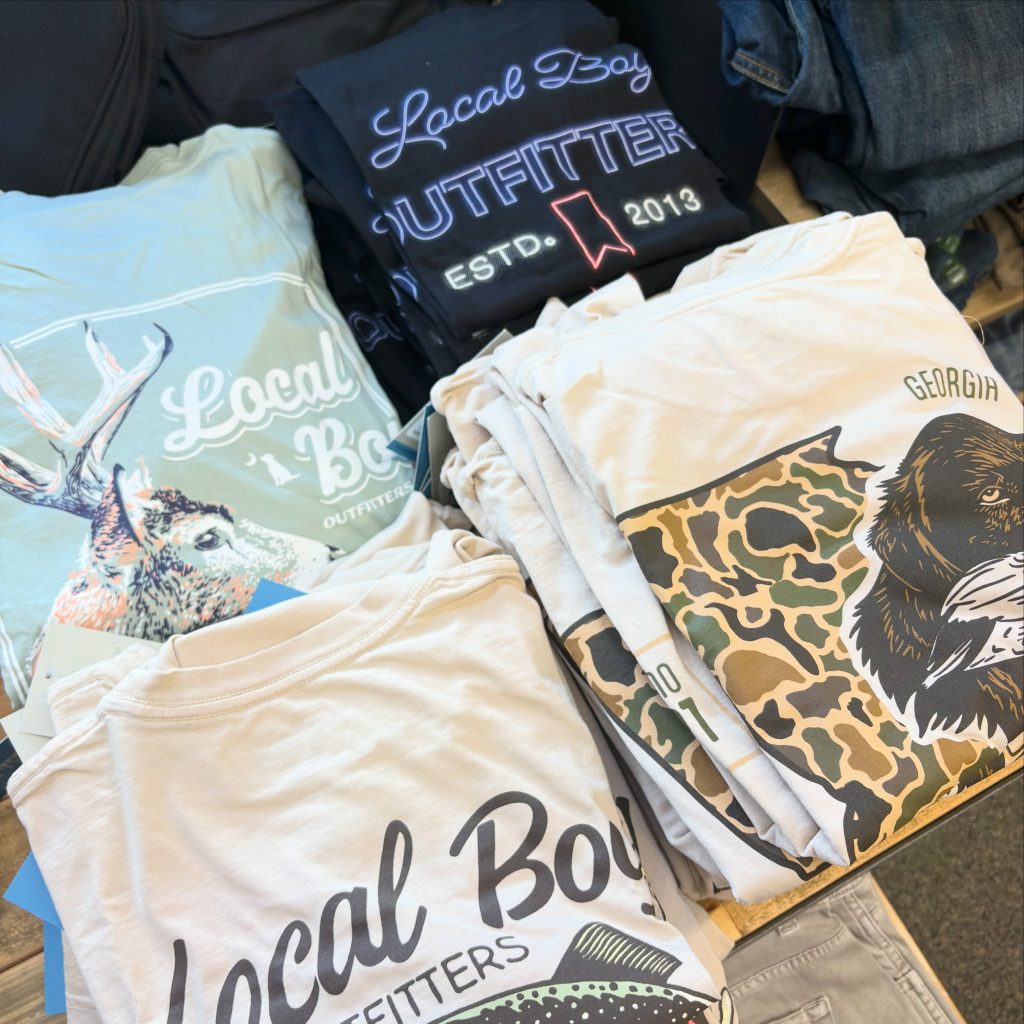 Pile of folded graphic t-shirts with various prints, including a dark shirt with neon-style lettering and a beige shirt showing 'GEORGIA' text visible at the edge.