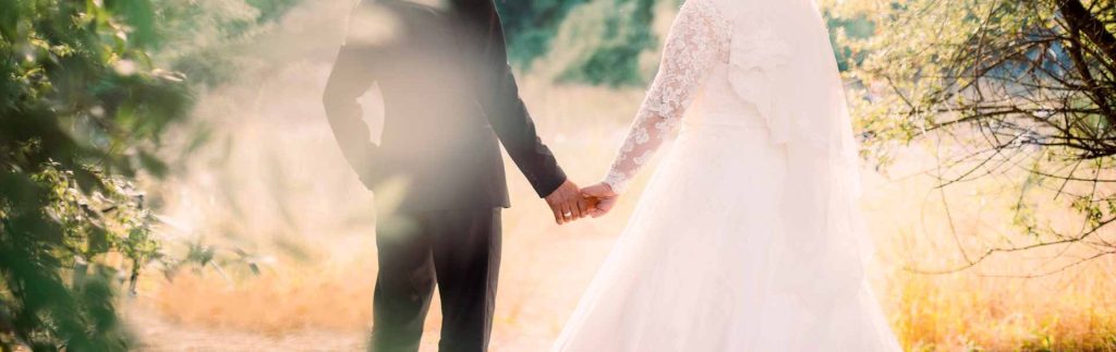 AdobeStock_222488229 - Reeves 1928 Bride and groom hold hands outdoors in warm, sunlit light; back view of wedding attire in a field setting.