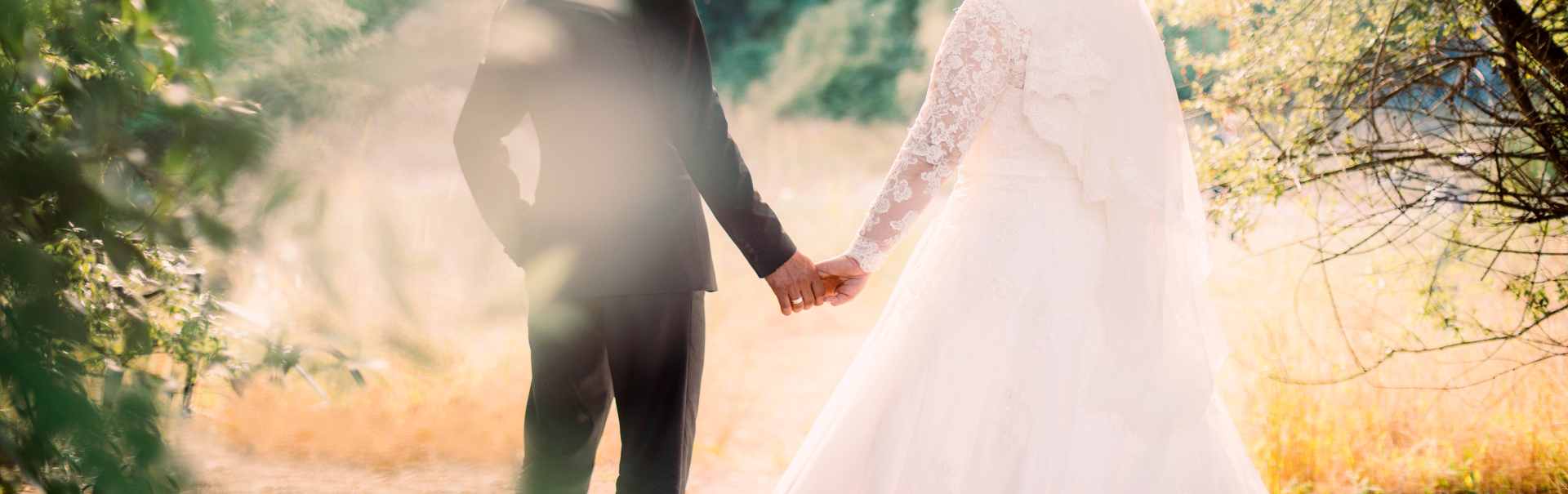 AdobeStock_222488229 - Reeves 1928 Bride and groom hold hands outdoors in warm, sunlit light; back view of wedding attire in a field setting.