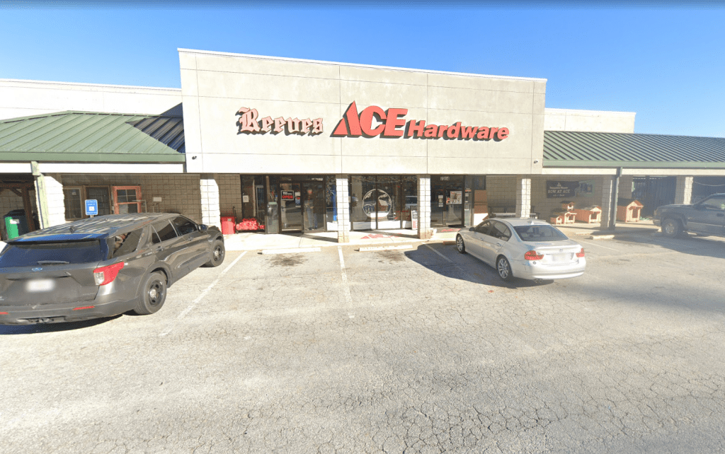 Front of an Ace Hardware store with two cars parked in a cracked parking lot and a clear blue sky above, storefront has stone columns and large glass doors.