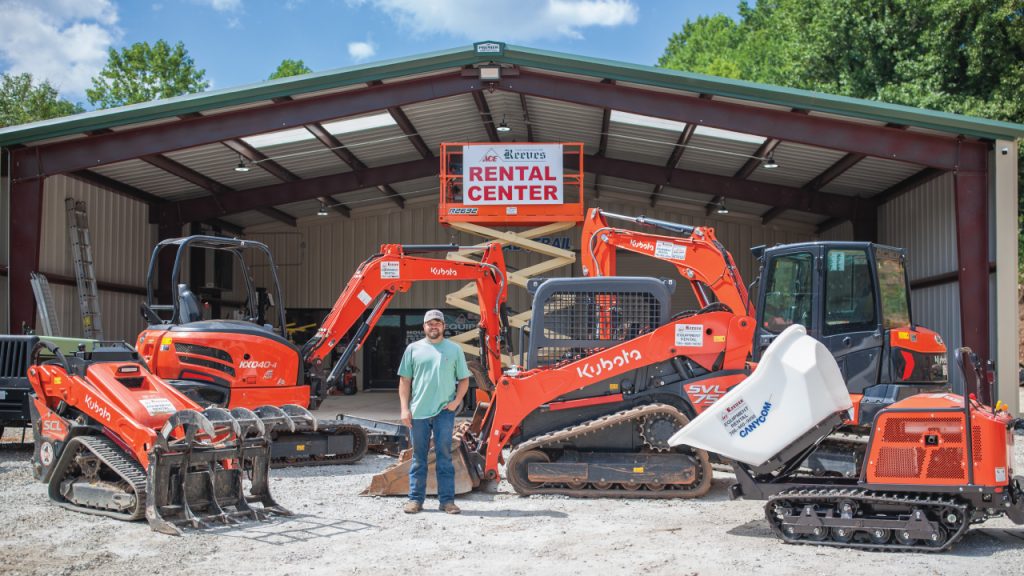 Man standing in front of orange Kubota excavators at a rental center under a large open-sided shed, sign reading 'Rental Center' above.