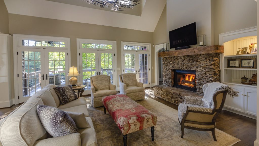 Cozy living room with a stone fireplace, beige sofa and chairs, and a red floral ottoman by the fire.
