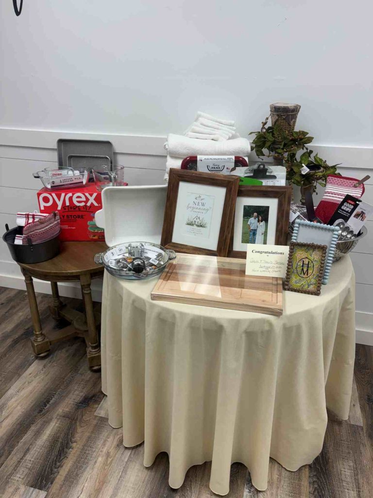Table display of gifts and keepsakes on a beige cloth: frames, folded towels, plant, gift items, and kitchenware including a Pyrex box.