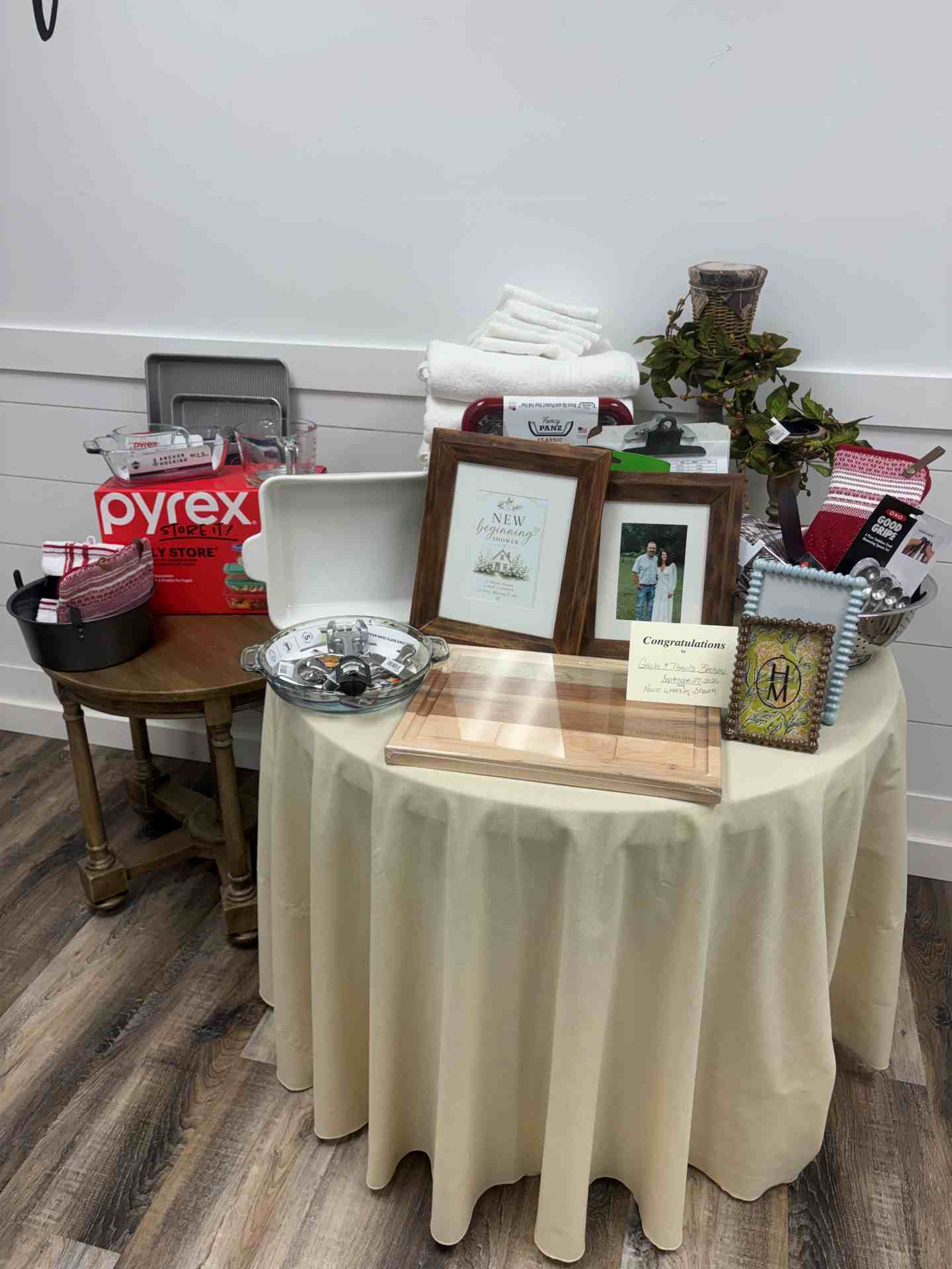 Table display of gifts and keepsakes on a beige cloth: frames, folded towels, plant, gift items, and kitchenware including a Pyrex box.