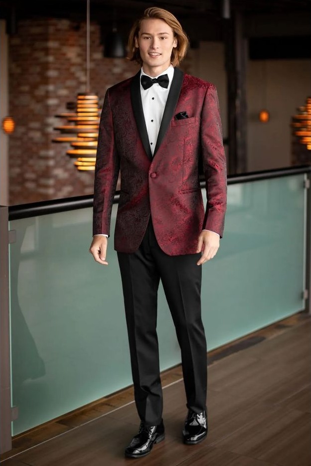 Reeves Tuxedo Rental (Apple Red Paisley Aries Tuxedo) - Reeves 1928 Young man in a burgundy velvet tuxedo jacket with black lapels, white shirt, black bow tie, standing indoors.