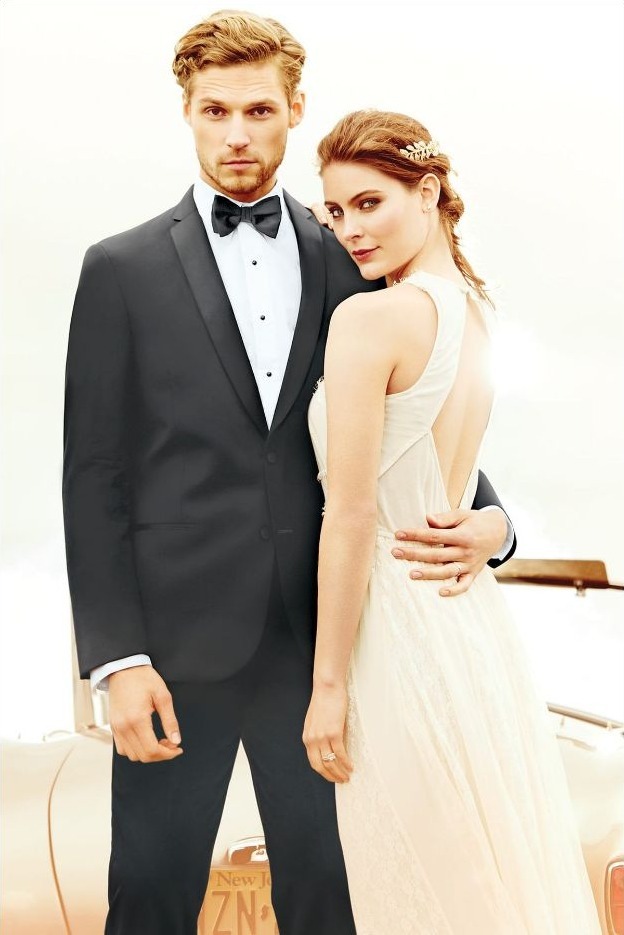 Reeves Tuxedo Rental (Black Berkeley Tuxedo) - Reeves 1928 Couple in formal wear posing together: man in a black tuxedo and bow tie, woman in an ivory gown with an open back, arm around him by a vintage car.
