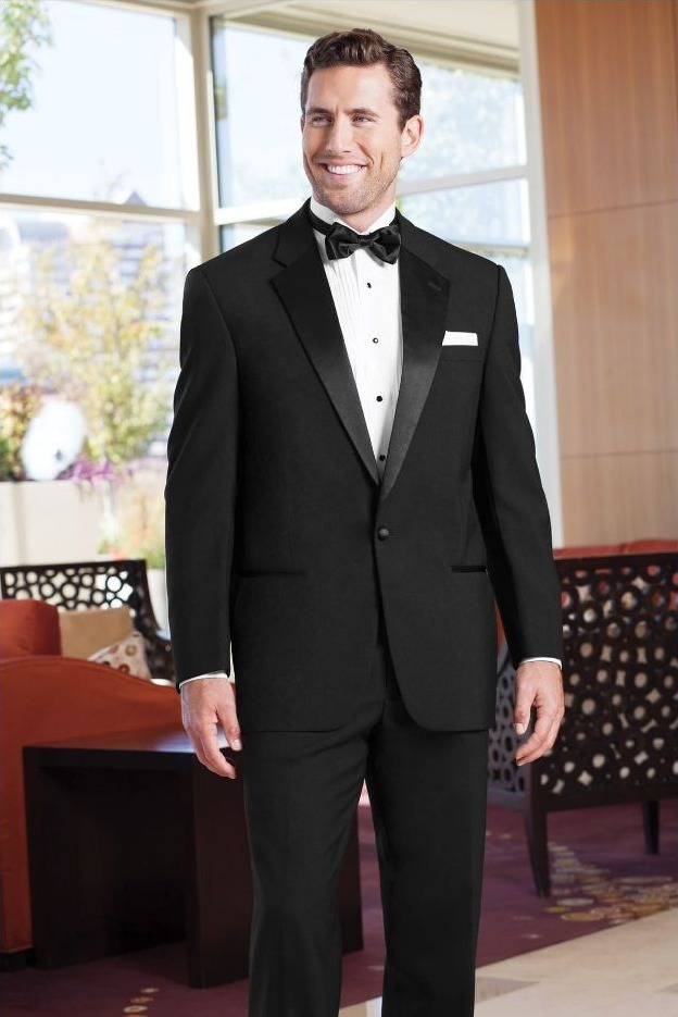 Reeves Tuxedo Rental (Black Classic Notch Tuxedo) - Reeves 1928 Smiling man in a black tuxedo with bow tie and white pocket square, standing in a bright lobby.