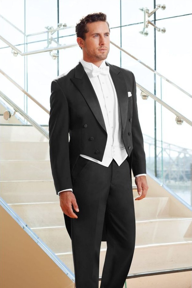Reeves Tuxedo Rental (Black Classic Peak Fulldress Tuxedo) - Reeves 1928 Man in a black tuxedo with a white vest and bow tie standing on a glass staircase in a modern lobby setting.