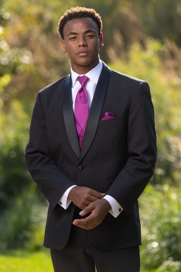 Reeves Tuxedo Rental (Black Cyprus Tuxedo) - Reeves 1928 Young man in a black suit with a magenta tie, standing outdoors with hands clasped.