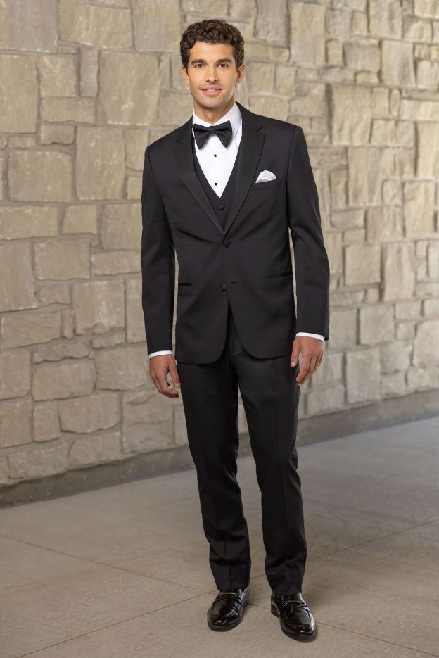 Reeves Tuxedo Rental (Black Force Performance Stretch Tuxedo) - Reeves 1928 Man in a black tuxedo with bow tie and pocket square standing against a stone wall.