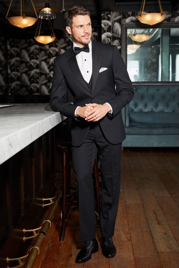 Reeves Tuxedo Rental (Black Legacy Performance Tuxedo) - Reeves 1928 Man in a black tuxedo with bow tie standing at a marble bar in a stylish lounge.
