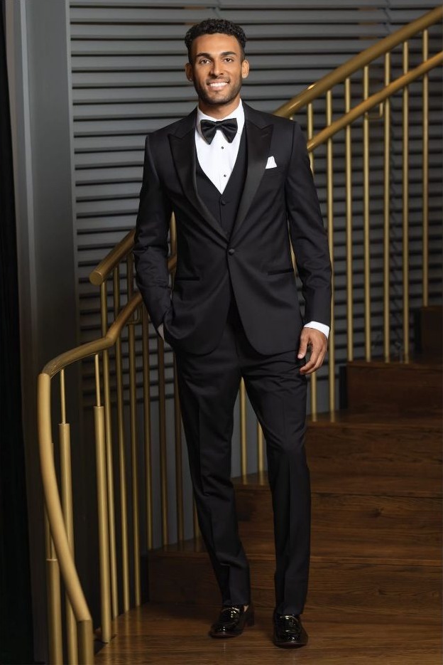 Reeves Tuxedo Rental (Black Milan Performance Tuxedo) - Reeves 1928 Man in a black tuxedo with bow tie standing on a wooden staircase, smiling at the camera, formal event attire.