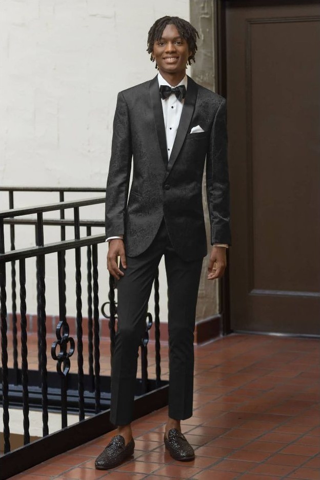 Reeves Tuxedo Rental (Black Paisley Aries Tuxedo) - Reeves 1928 Young man in a black tuxedo with bow tie and pocket square standing on a tiled balcony.