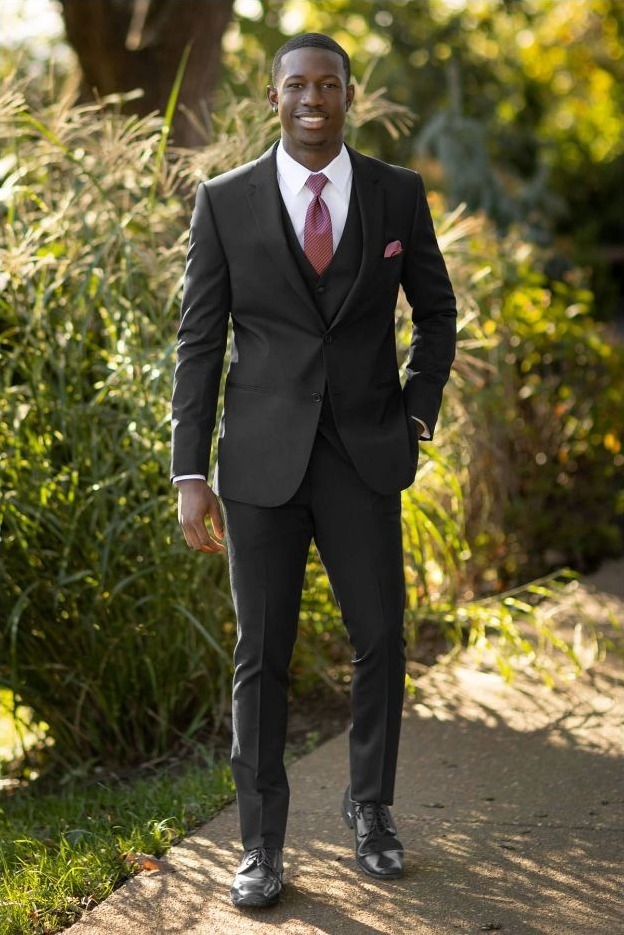 Reeves Tuxedo Rental (Black Performance Wedding Suit) - Reeves 1928 Man in a black suit with a red tie and pocket square stands on a sunny garden path.