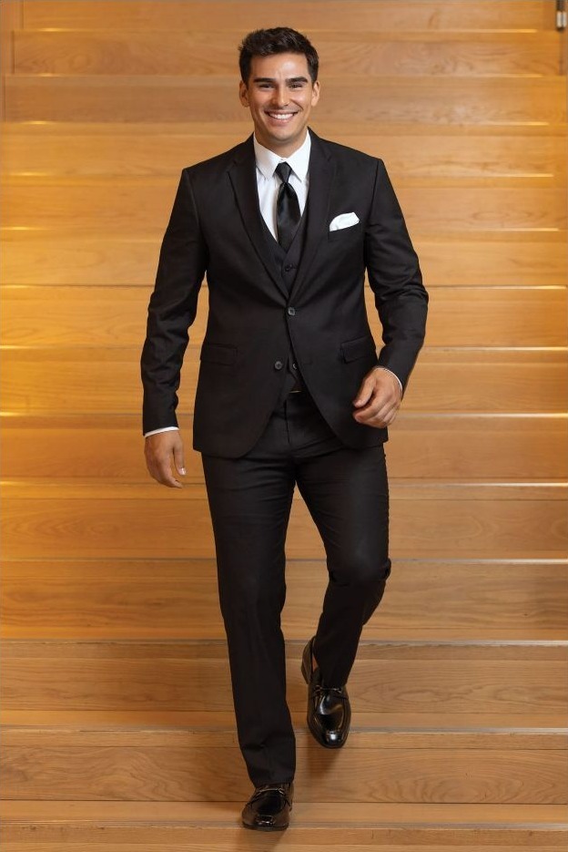 Reeves Tuxedo Rental (Black Stretch Suit) - Reeves 1928 Man in a black suit with white shirt and tie walking down wooden stairs, smiling at the camera.