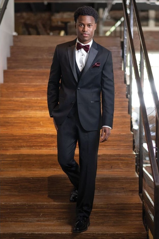 Reeves Tuxedo Rental (Black Stretch Tuxedo) - Reeves 1928 Man in a black tuxedo with burgundy bow tie walking down wooden stairs indoors, posing confidently.