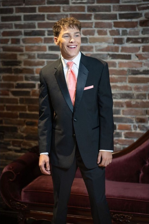 Reeves Tuxedo Rental (Black Troy Tuxedo) - Reeves 1928 Man in a dark suit with pink tie and pocket square, smiling against a brick wall background