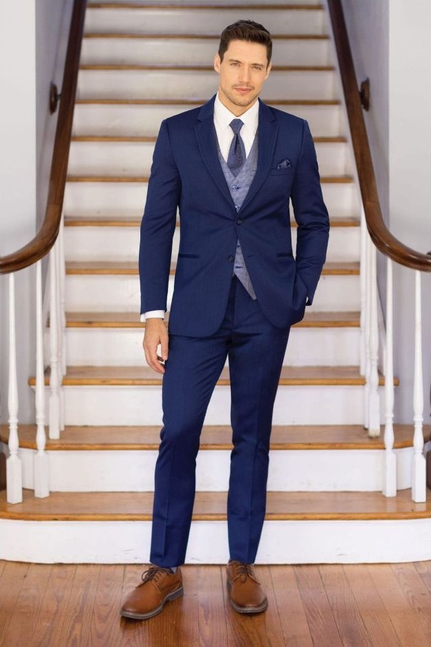 Reeves Tuxedo Rental (Blue Performance Wedding Suit) - Reeves 1928 Man in a navy suit with a vest and tie standing on a wooden staircase.