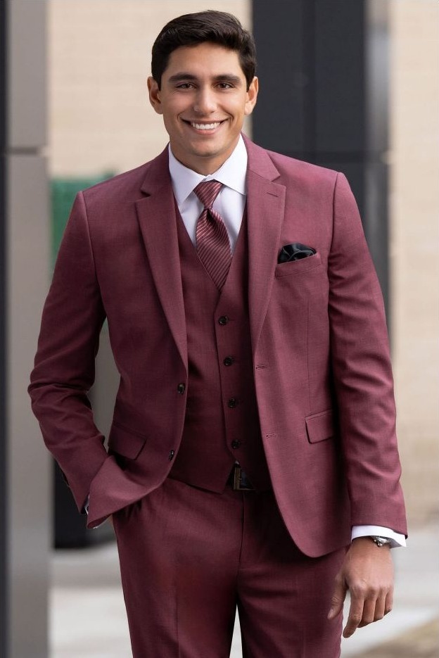 Reeves Tuxedo Rental (Brick Stretch Suit) - Reeves 1928 Man in a burgundy suit with a vest and pocket square, smiling outdoors and standing with one hand in his pocket.