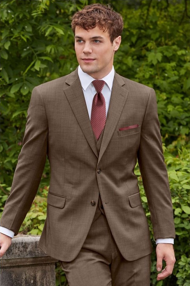 Reeves Tuxedo Rental (Brown Archer Slim Fit Suit) - Reeves 1928 Young man in a brown suit with a white shirt and burgundy tie, standing outdoors with greenery in the background.