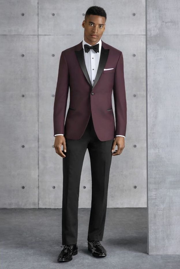 Reeves Tuxedo Rental (Burgundy Empire Tuxedo) - Reeves 1928 Man in a burgundy tuxedo with black lapels, white shirt, black bow tie, and black trousers standing against a concrete wall.