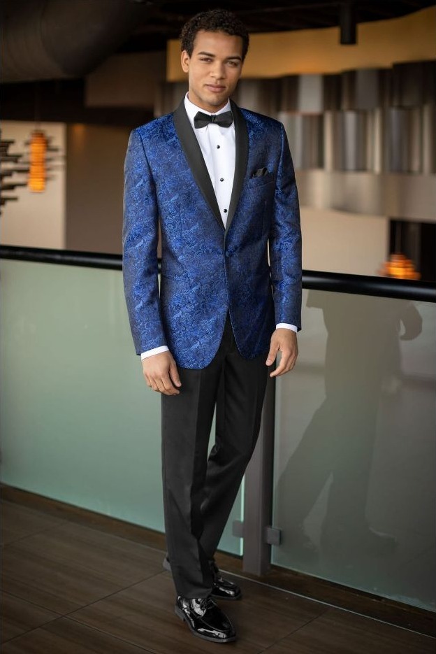 Reeves Tuxedo Rental (Cobalt Blue Paisley Aries Tuxedo) - Reeves 1928 Young man in a blue patterned tuxedo jacket, white dress shirt, black bow tie, and black trousers posing indoors.