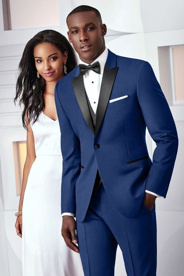 Reeves Tuxedo Rental (Cobalt Blue Tribeca Tuxedo) - Reeves 1928 Man in a blue tuxedo with black lapels and a bow tie poses beside a woman in a white gown in a bright, modern interior.