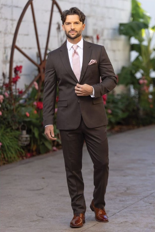 Reeves Tuxedo Rental (Dark Brown Stretch Suit) - Reeves 1928 Man in a brown suit with pink tie walking outdoors, wearing brown dress shoes and a light pocket square in a garden setting