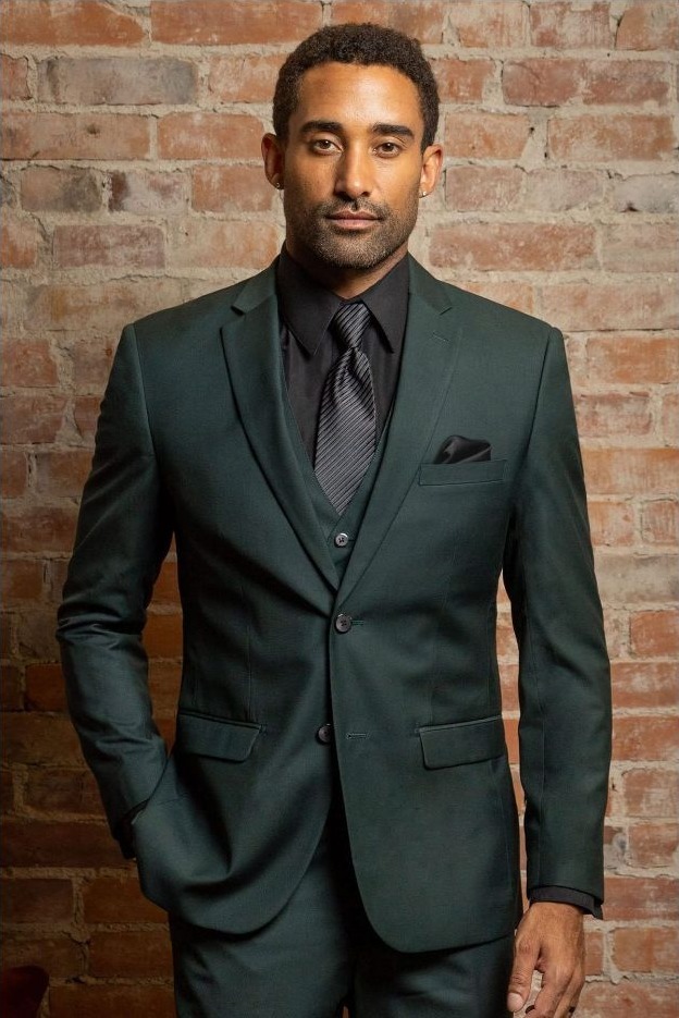 Reeves Tuxedo Rental (Dark Green Stretch Suit) - Reeves 1928 Man in a dark green suit with black shirt and tie, standing against a brick wall, with a pocket square in the jacket pocket.
