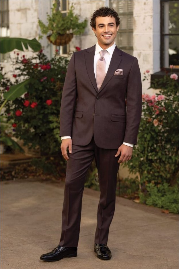 Reeves Tuxedo Rental (Eggplant Stretch Suit) - Reeves 1928 Smiling man in a dark brown suit with pink tie and pocket square standing outdoors among flowers and a stone building.