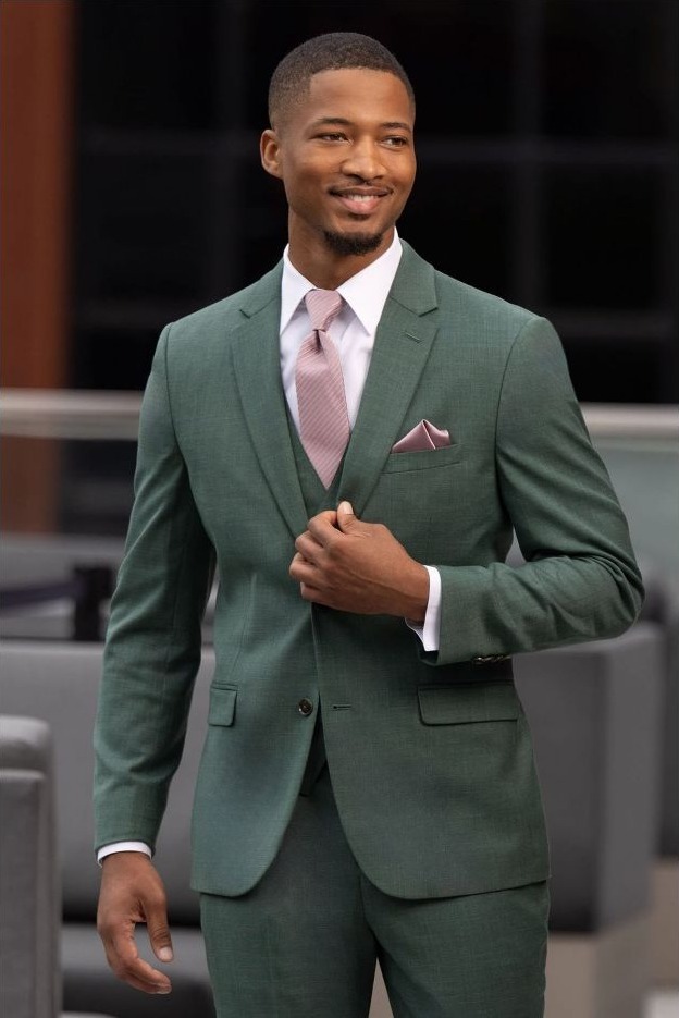 Reeves Tuxedo Rental (Forest Green Stretch Suit) - Reeves 1928 Man in a green suit adjusts his jacket, wearing a pink tie and pocket square, smiling confidently.