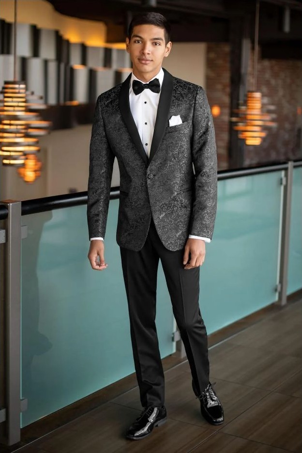 Reeves Tuxedo Rental (Granite Paisley Aries Tuxedo) - Reeves 1928 Young man in a patterned black tuxedo with a bow tie, white shirt, and pocket square in a stylish indoor venue.