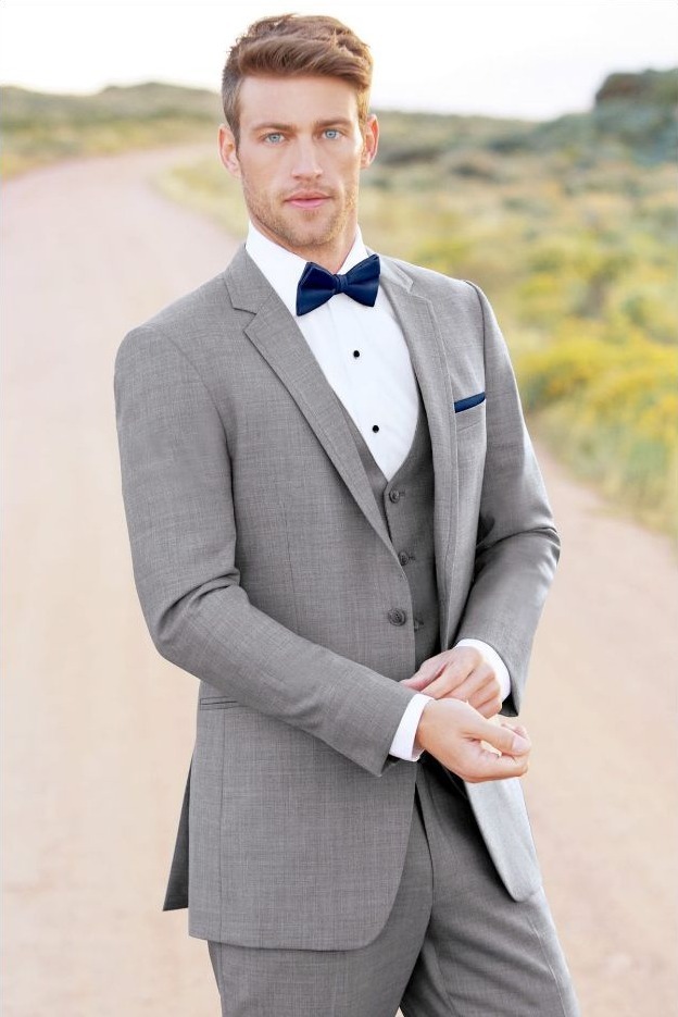 Reeves Tuxedo Rental (Heather Grey Clayton Suit) - Reeves 1928 Man in a light gray suit with a navy bow tie, white shirt, and vest, standing on a dirt road outdoors with blurred countryside behind him.