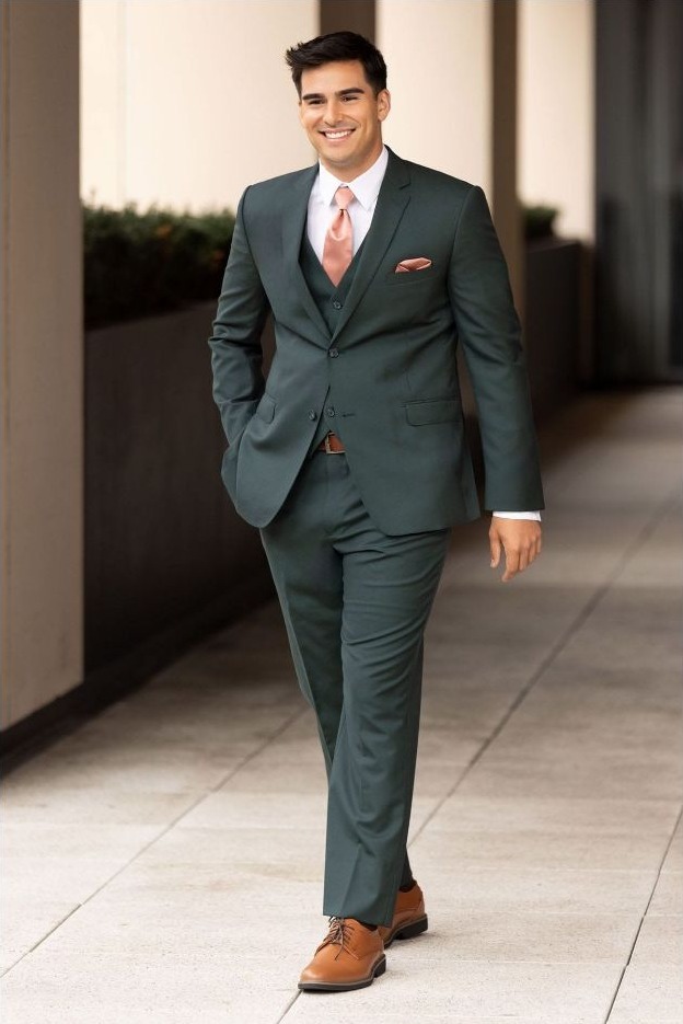 Reeves Tuxedo Rental (Hunter Green Luka Suit) - Reeves 1928 Smiling man in a teal-green suit, pink tie, and brown shoes walking outdoors along a sidewalk.