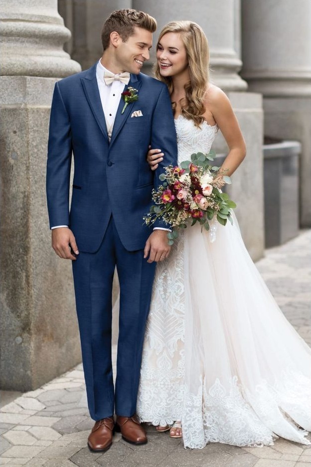 Reeves Tuxedo Rental (Indigo Blue Lane Tuxedo) - Reeves 1928 Bride in a white lace gown and groom in a navy suit smile as they stand outdoors, holding hands with a floral bouquet nearby.