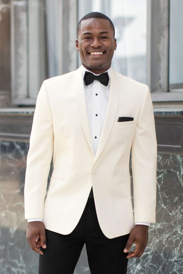 Reeves Tuxedo Rental (Ivory Classic Shawl Tuxedo) - Reeves 1928 Man in a cream tuxedo with black bow tie, white shirt, and black pocket square, smiling outdoors in front of a building.