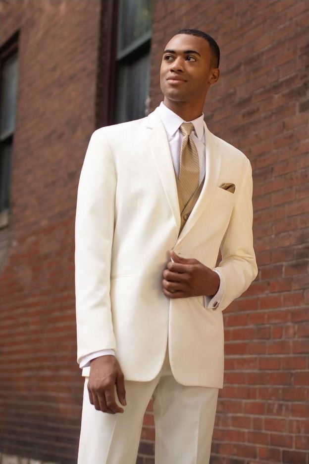 Reeves Tuxedo Rental (Ivory Troy Tuxedo) - Reeves 1928 Man in a cream suit adjusting his jacket against a brick wall, wearing a light beige tie and pocket square.